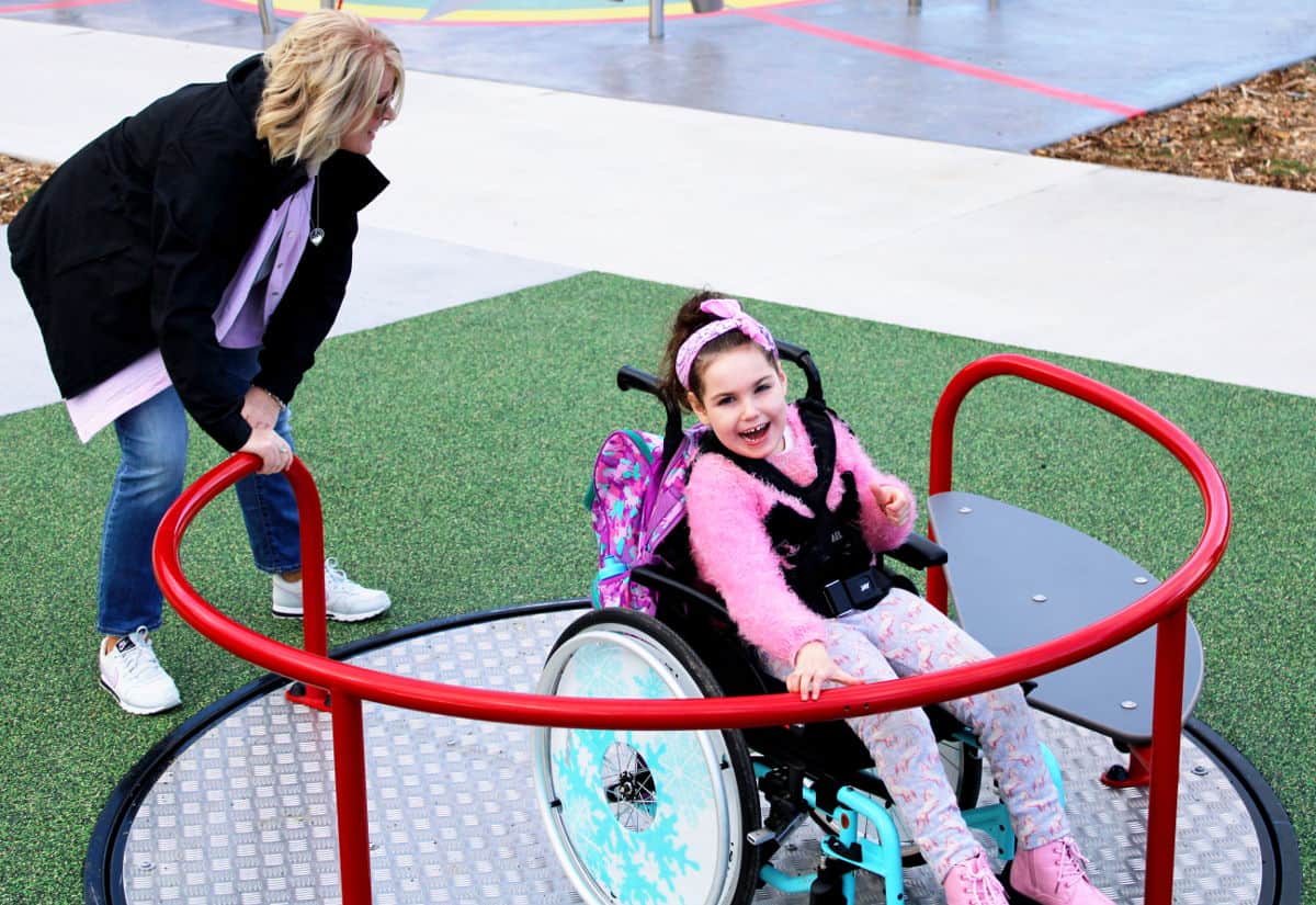 Smiling girl in a wheelchair using the accessible carousel at Maidstone Max Playground with caregiver assistance | Photo: Playground People Ltd
