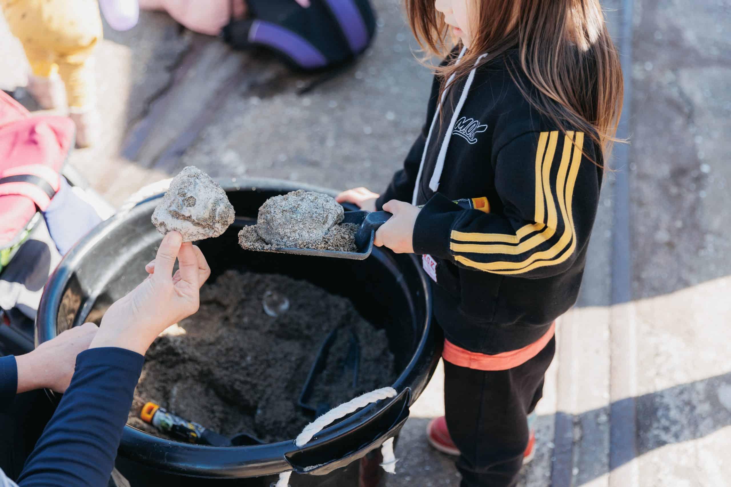 Child holding a small spade and digging through sand in a large bucket while another person holds up a piece of fossil-like rock at a hands-on outdoor learning activity.