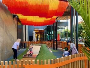 Small children's playground at Sylvia Park with a hill slide, orange and yellow canopy, and seating nearby.