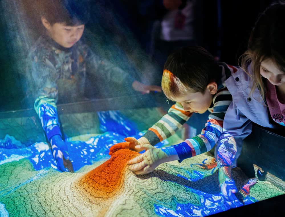 Children using an interactive augmented reality sand table to learn about landforms and weather at MOTAT