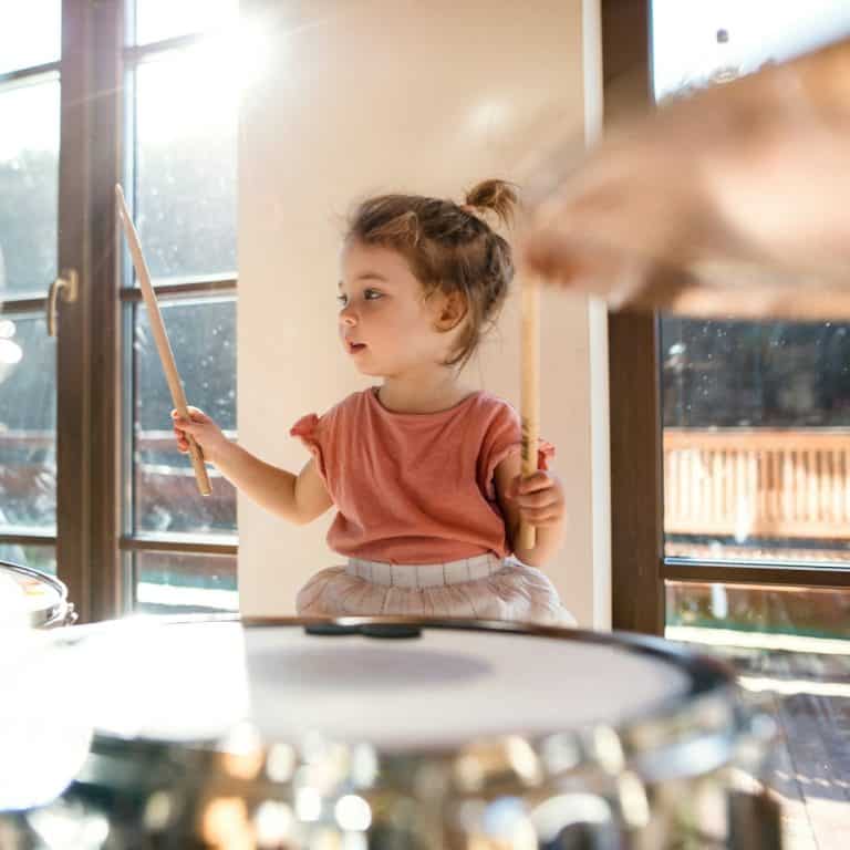Young child enthusiastically playing a drum kit with sticks in a sunlit room