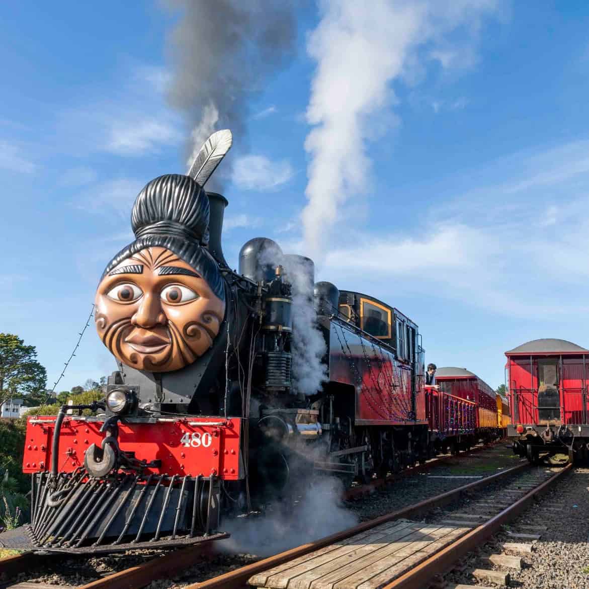 Vintage steam train decorated with a Māori face design on the front and plume of smoke in the air