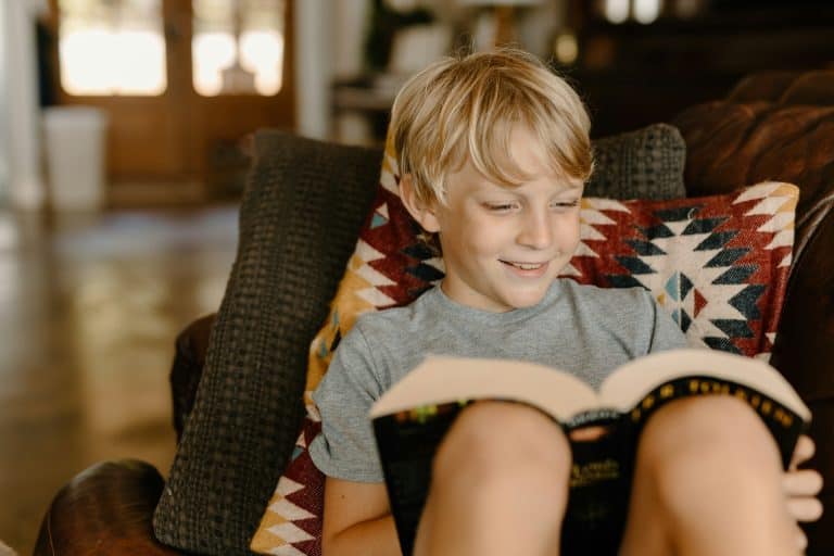 Young boy smiling while reading a book on the couch at home.
