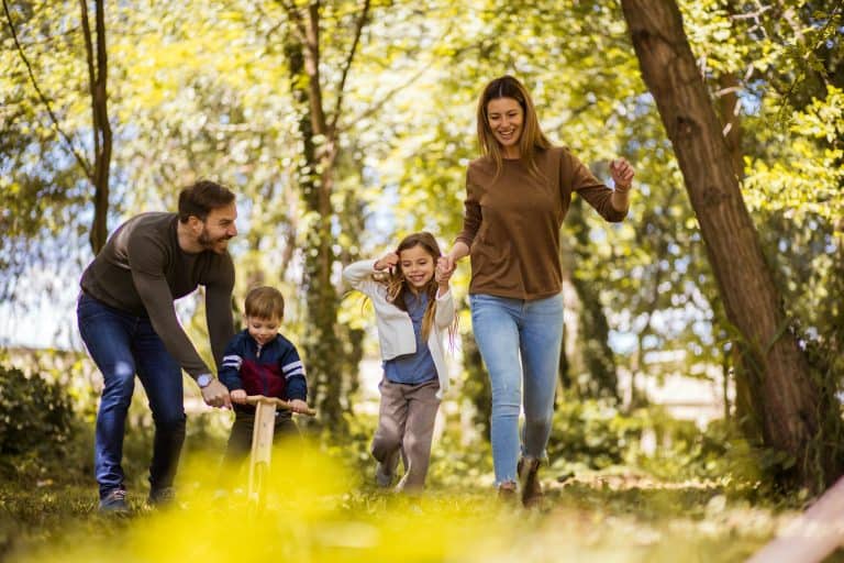 Family with two young children walking and riding a balance bike through a leafy park.