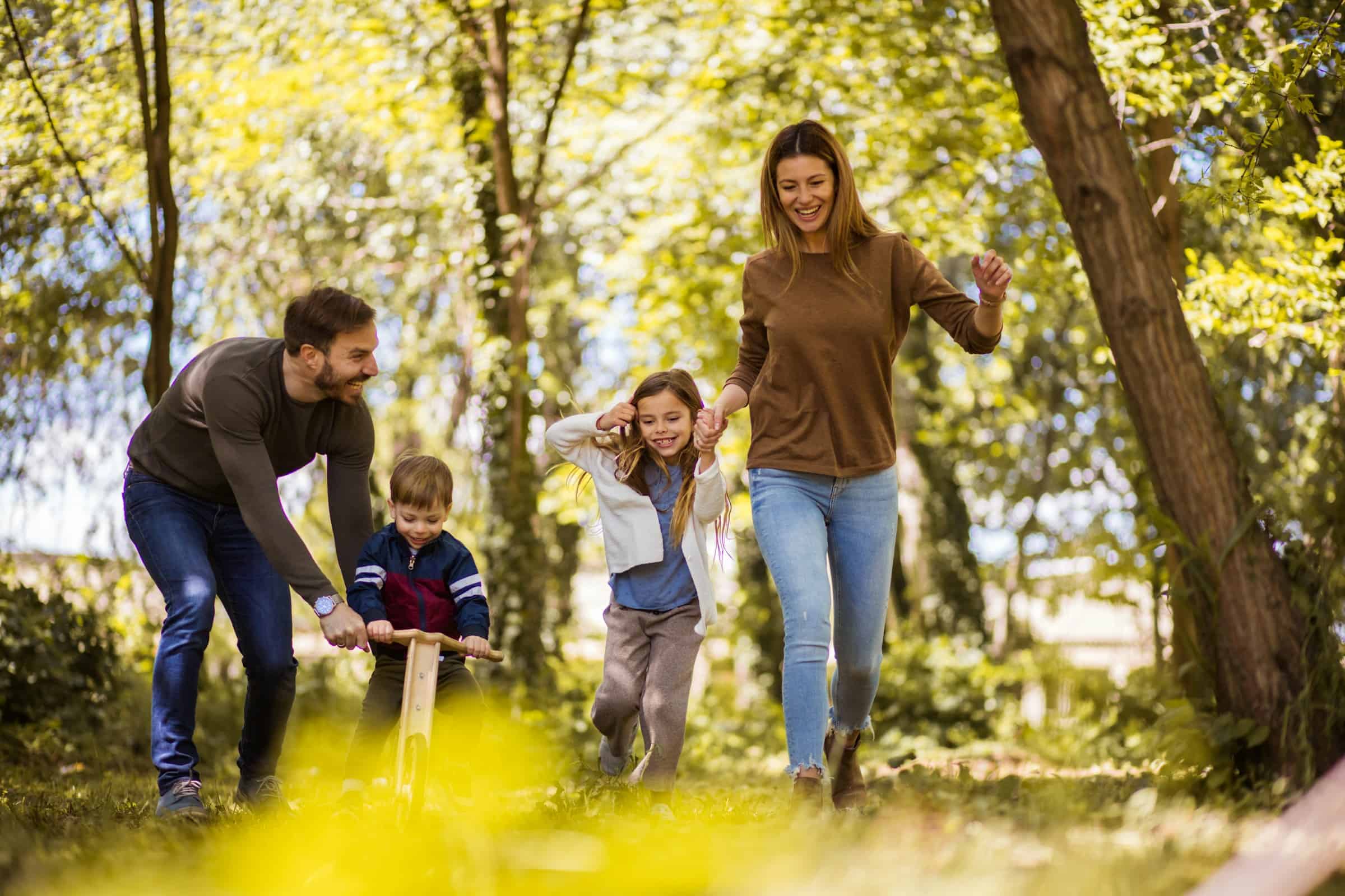 Family with two young children walking and riding a balance bike through a leafy park.