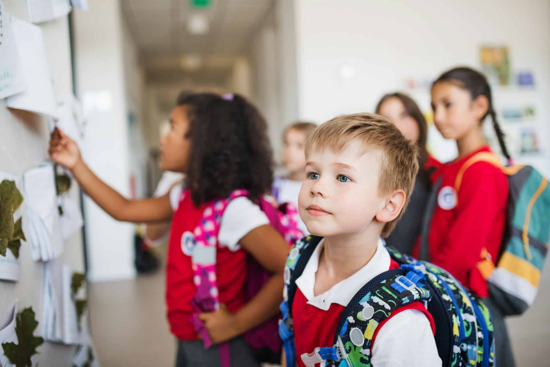 Primary school children in uniform with backpacks looking at a wall display inside a school corridor.