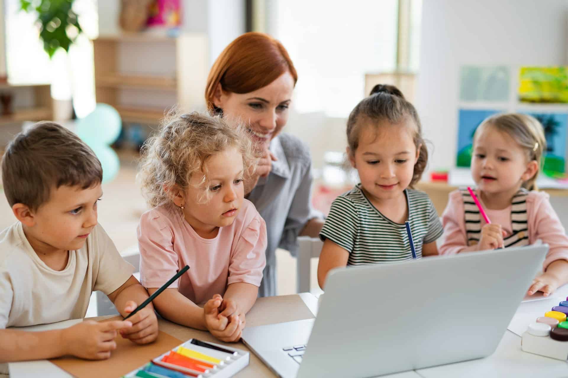 Young children and a teacher gathered around a laptop in a classroom, holding coloured pencils.