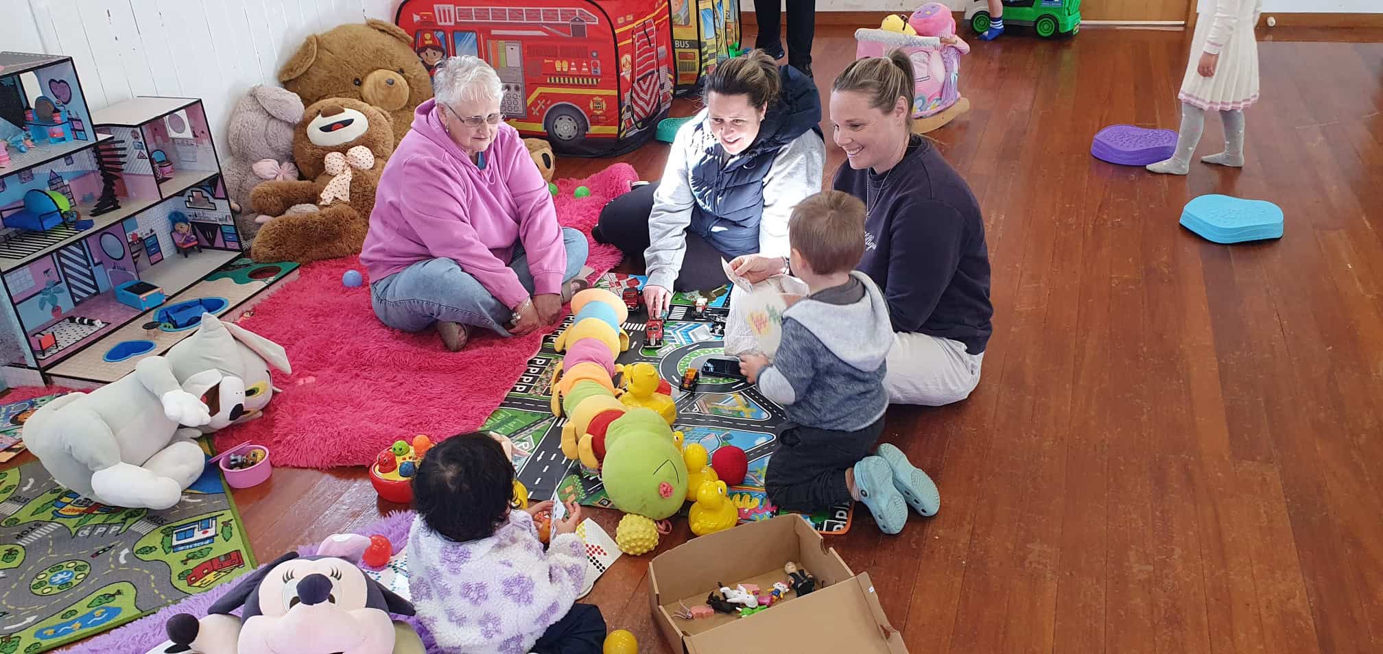 Adults and young children playing together indoors with soft toys, cars and games on colourful mats during a preschool play session.